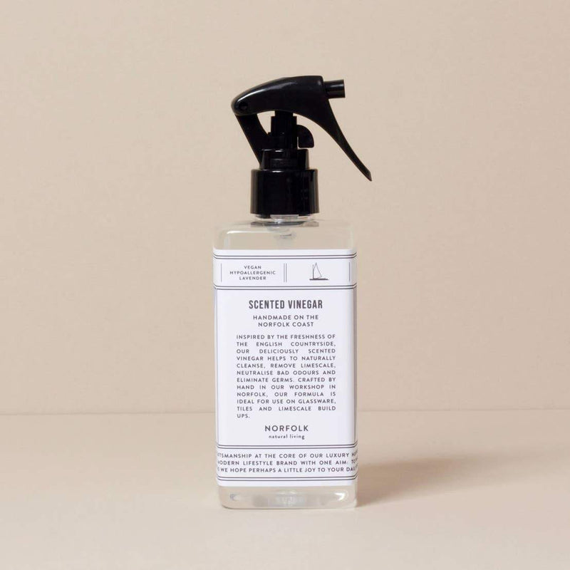 A clear spray bottle of Norfolk Natural Living Lavender Scented Vinegar, enhanced with essential oils, stands against a plain beige background. The label details its ingredients and uses in elegant typesetting as a natural cleaning product.