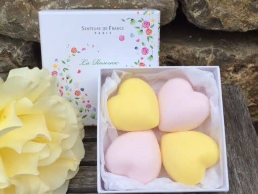 An open box containing three Senteurs De France Coffret “Roseraie” Honeysuckle & Rose heart-shaped soaps in pastel pink and yellow, alongside a blooming yellow flower and a product box labeled "senteurs de france," placed on a rustic stone.