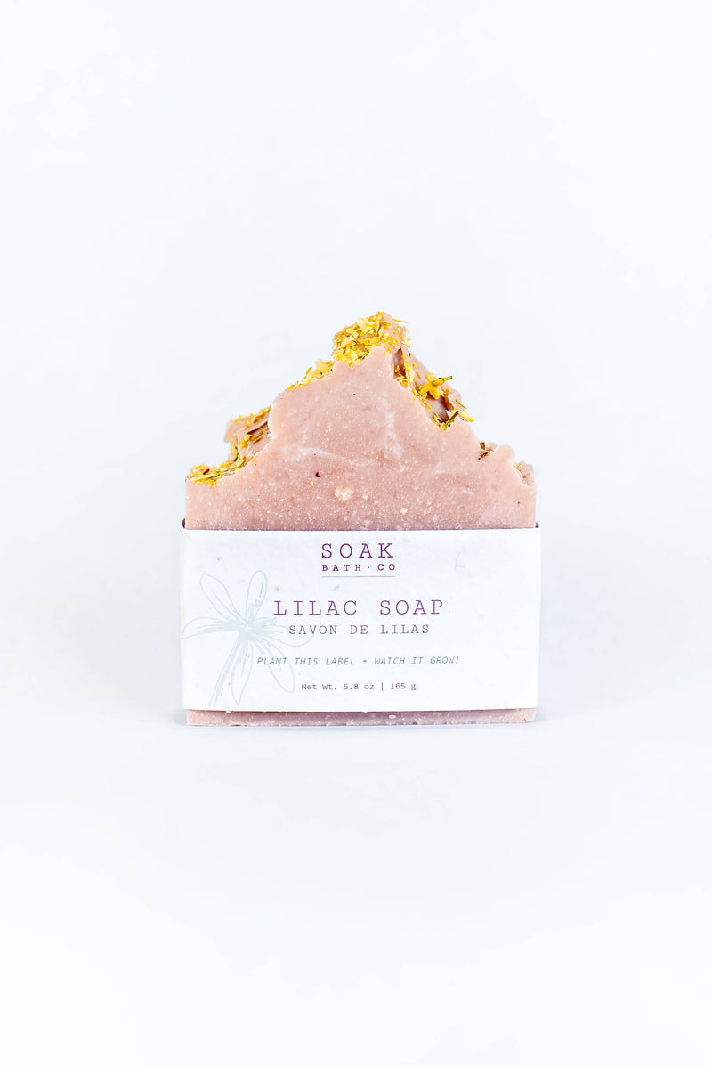 A bar of SOAK Bath Co. - Lilac Soap Bar topped with gold leaf, wrapped in a labeled white paper band against a bright white background.