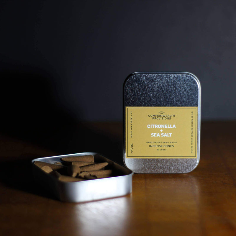 A metal tin labeled "Commonwealth Provisions Citronella + Sea Salt Incense Cones" with its lid removed to reveal several brown square blocks inside, resting on a dark wooden surface against a shadowy background.
