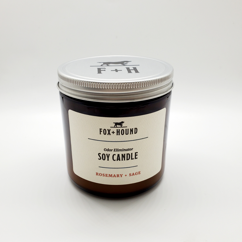 A soy candle in a clear glass jar labeled "Fox + Hound Odor Eliminator Soy Candle - Rosemary + Sage" on a white background.