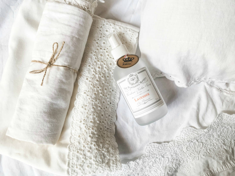 A Z&Co. frosted glass bottle of Lavender Farmhouse room/linen spray lying beside white lace and linen items on a soft, textured background, conveying a clean and serene aesthetic.