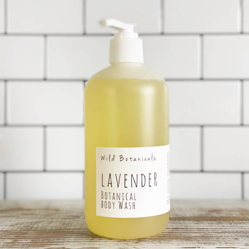 A large pump bottle of Wild Botanicals Lavender Botanical Body Wash stands in front of a white brick background. The liquid inside the transparent bottle is yellowish.