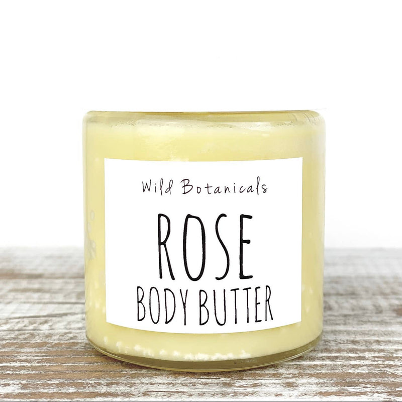 A jar of Wild Botanicals - Rose Body Butter with a creamy, textured appearance, labeled clearly in black script on a white background, set against a light wooden surface. Enhanced with organic shea.