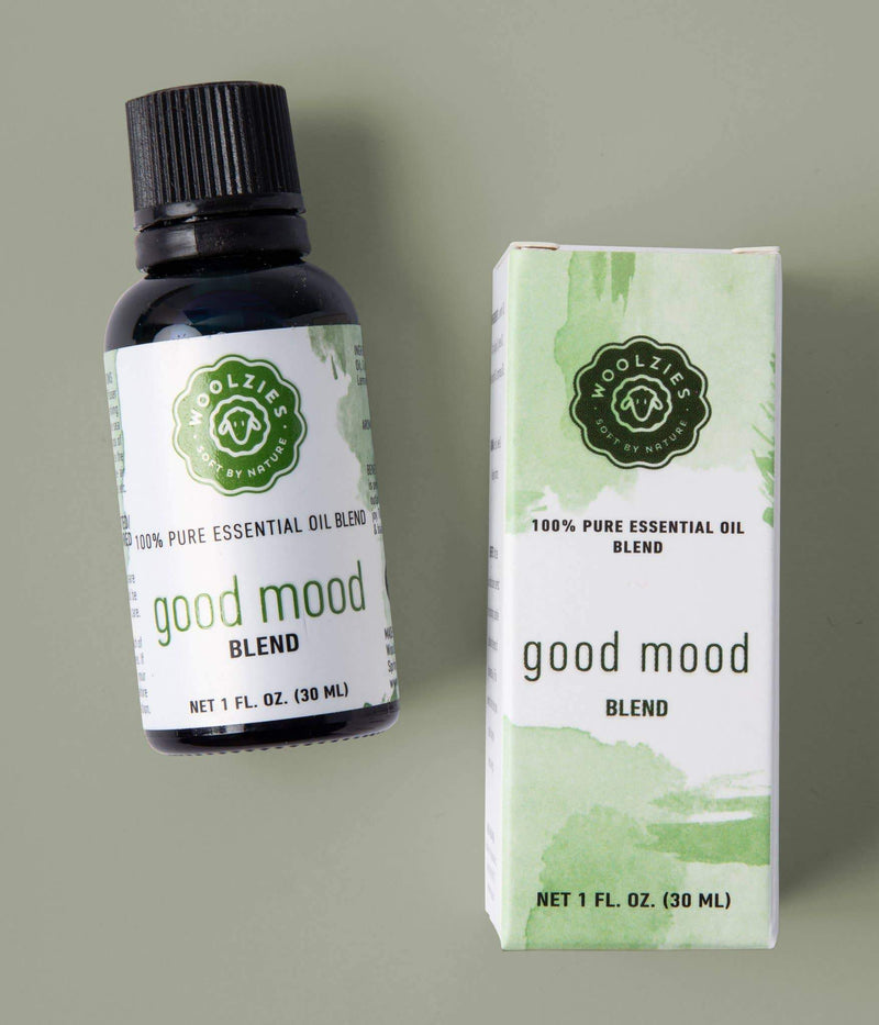 A bottle of Woolzies Good Mood Blend Essential Oil next to its packaging box, both labeled with green watercolor design. The box and bottle display product details and are placed against a light green background.