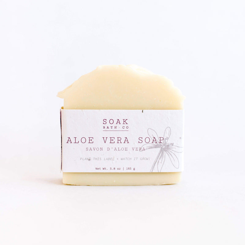 A bar of SOAK Bath Co. - Aloe Vera Soap in zero waste, plantable seed paper packaging with a sketch of an aloe plant. The label features text and the soap has a w