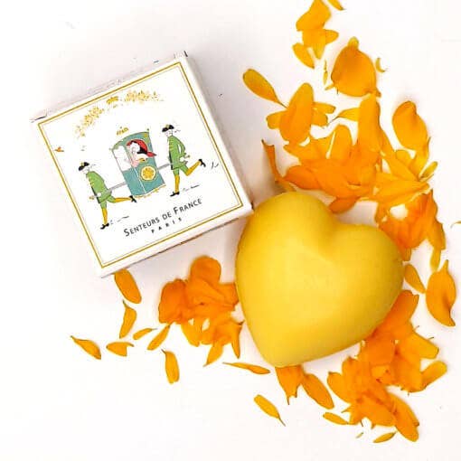 A Senteurs De France Honeysuckle Heart Soap inspired by Marie-Antoinette next to a perfume box with a vintage French illustration, surrounded by scattered orange flower petals on a white background.