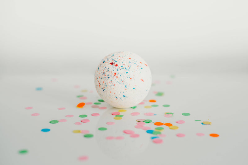 A speckled white egg stands at the center on a reflective white surface, surrounded by scattered colorful confetti resembling a SOAK Bath Co. - Birthday Cake bath bomb.