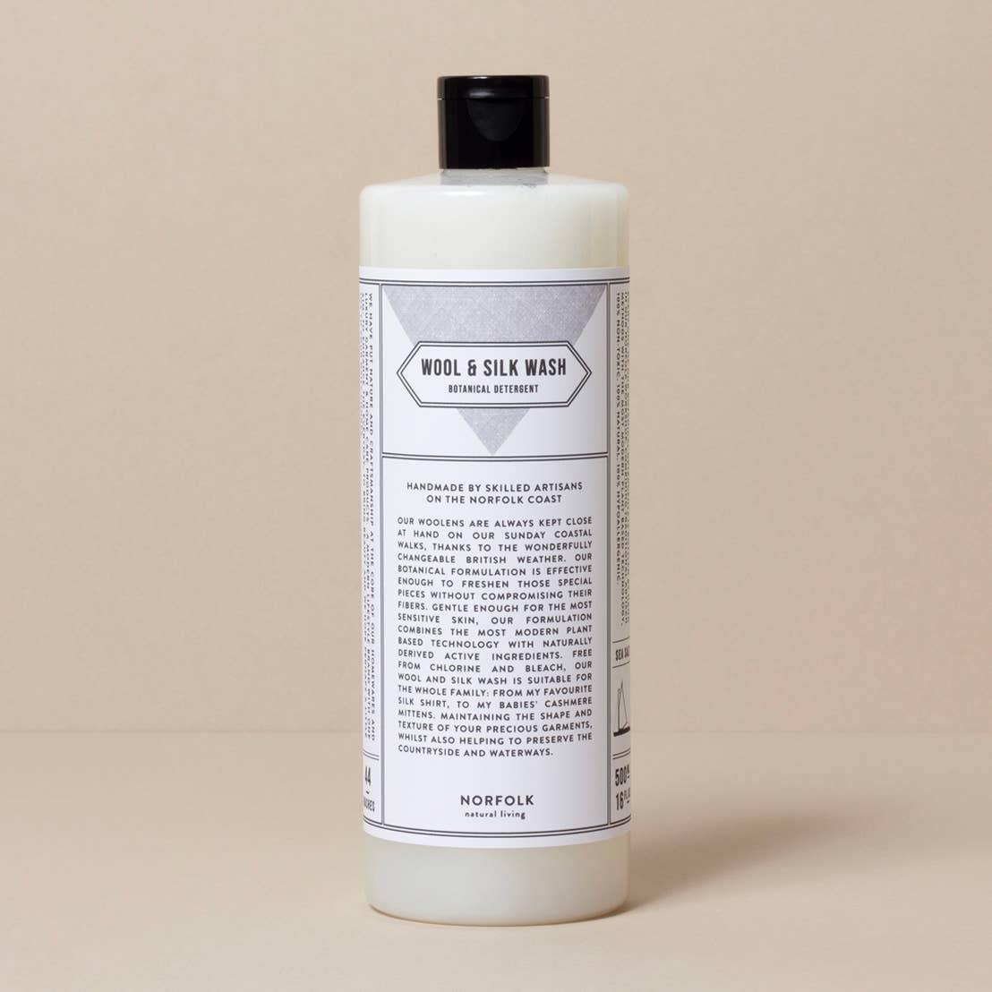 A white bottle of Norfolk Natural Living Coastal Wool and Silk Detergent with a black cap on a beige background. The label, featuring gentle formulation details, provides extensive product and usage information.
