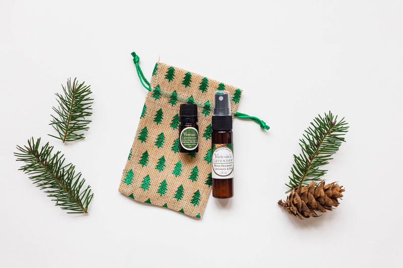 Two bottles of Victoria's Lavender - Lavender & Fir Essential Oil next to a burlap pouch with a pine tree pattern, arranged on a white background along with pine branches and a pine cone.