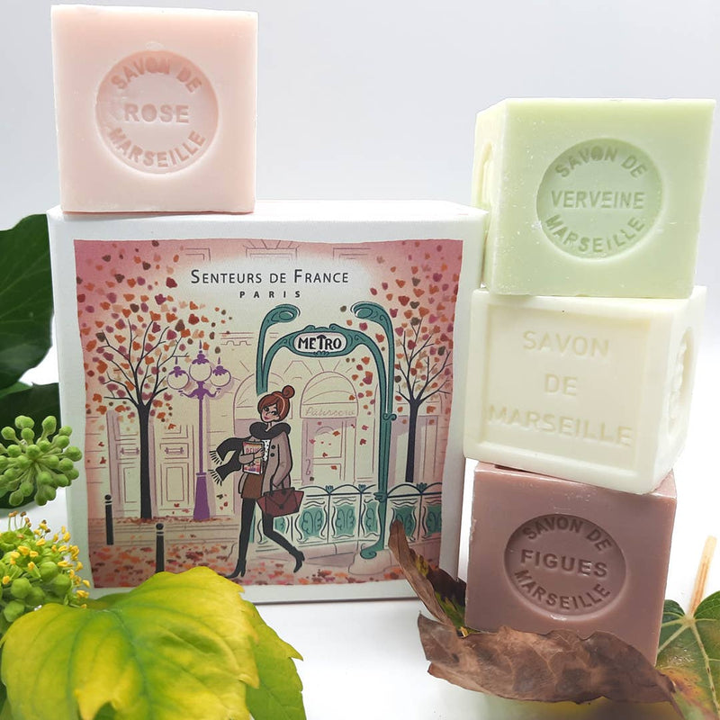 Assorted bars of Senteurs De France cube soaps in rose, verbena, and fig scents, displayed with an illustrated Paris-themed box featuring a person by a metro entrance. Leaves and flowers accent the setup.