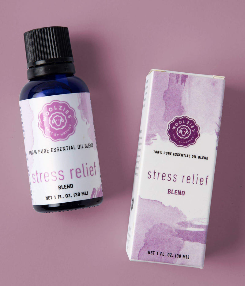 A blue glass bottle labeled "100% pure Woolzies Stress Relief Blend Essential Oil" next to its white cardboard packaging against a pink background. Both items feature purple Woolzies accents and branding logos.