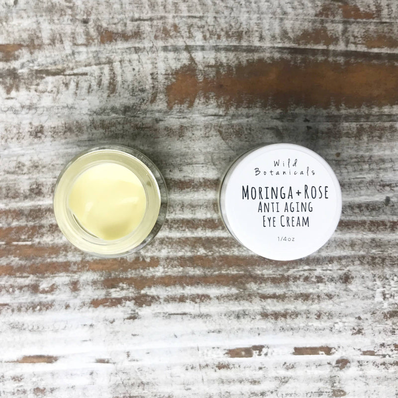 Overhead view of an open jar of Wild Botanicals - Moringa and Rose Anti Aging Eye Cream next to its lid, both on a wooden surface. The jar is half-filled with a yellowish cream.