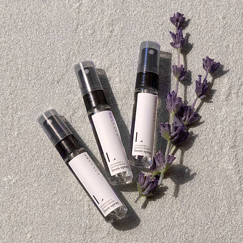 Three bottles of Lavande Lavender-Sandalwood Mini Linen Spray ( 5ml ) with lavender sprigs on a textured gray background, arranged diagonally with sunlight casting soft shadows.