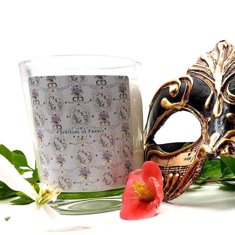 A Senteurs De France Candle Versailles Lavender with a decorative floral pattern on its holder, next to an elaborate gold and black carnival mask, set against a white backdrop with green.