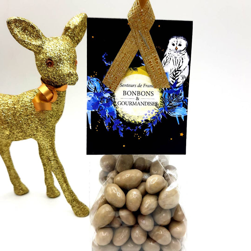 A golden glittery reindeer figurine next to a Senteurs De France Noël Chocolate Raisins Confectionery with Champagne Marc featuring an artistic owl design, with a container of almond paste in the foreground. White background.