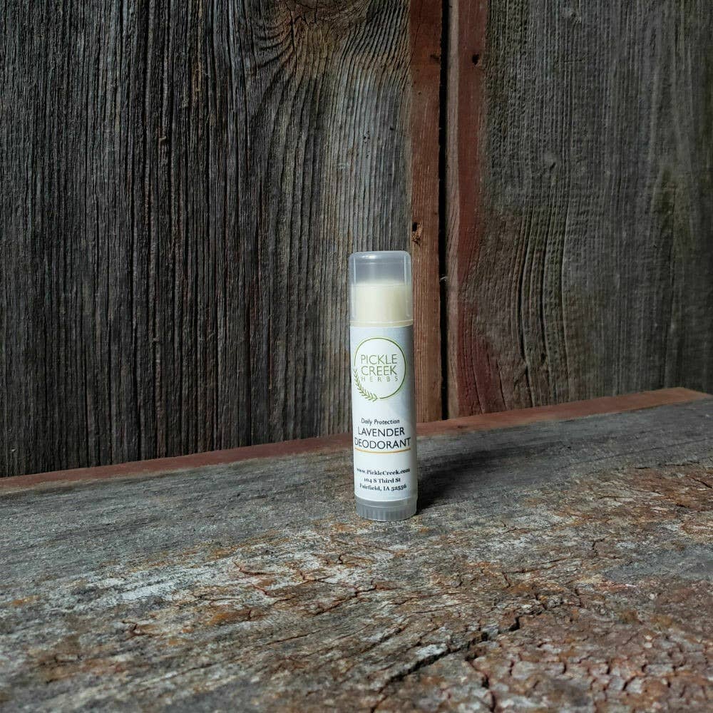 A stick of Pickle Creek Herbs Lavender Deodorant for Daily Protection, resting on a wooden surface against a textured wood backdrop, presented in a simple, cylindrical container. This natural deodorant is paraben-free.