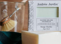 A close-up of Andrée Jardin Solid Dish Washing Soap Sage Basil labeled "sauge basilic" beside a large yellow soap block and a wooden dish brush in blurred background.
