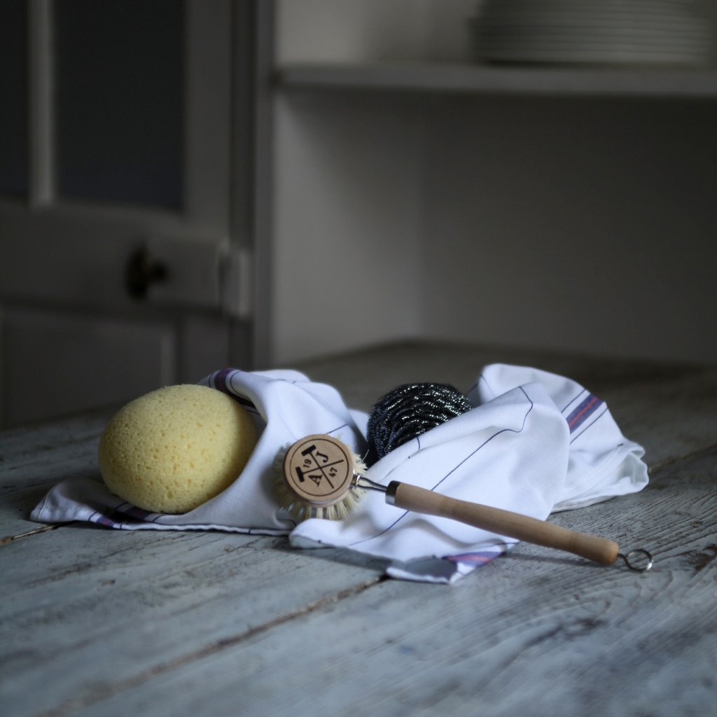 A kitchen towel with a natural sponge, a black scrubber, and an Andrée Jardin Tradition Handled Dish Brush with a peace symbol rests on a rustic wooden table.