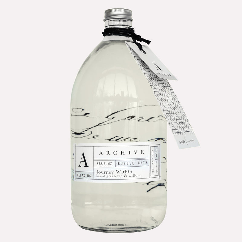 A large clear glass bottle labeled "Archive by Margot Elena Journey Within Bubble Bath," containing a luxurious soak with a tag and sketch of a building attached, on a white background.