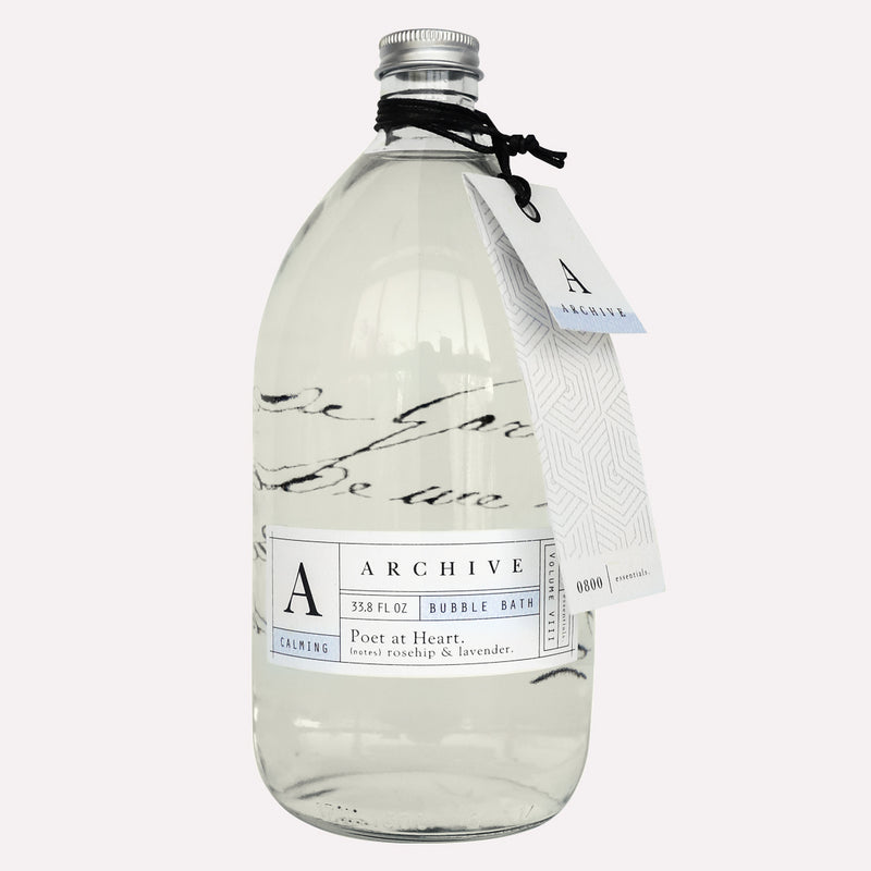 Clear glass bottle of Archive by Margot Elena Poet at Heart Bubble Bath with elegant labeling, filled with a transparent liquid and sealed with a black cap, accompanied by a hanging white tag.