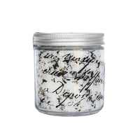 A clear glass jar filled with shredded paper, featuring black handwritten text, sealed with a metal lid. The background is plain white, emphasizing the jar's contents infused with purifying minerals of the Margot Elena ARCHIVE Still Waters Salt Soak.