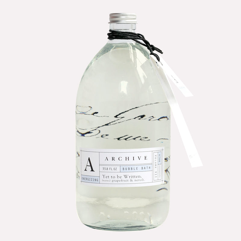 Clear plastic bottle containing a light-colored liquid, labeled as "Archive by Margot Elena Yet To Be Written Bubble Bath" with grapefruit and neroli scents. This transformative blend includes natural oils. The bottle has a white cap and decorative design.