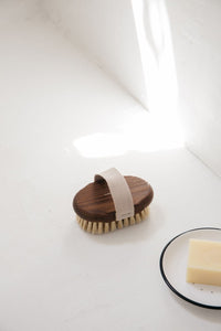 An Andrée Jardin Tradition Ash Wood Massage Brush with a strap, placed upright on a white surface beside a bar of soap on a black-rimmed dish, lit by natural light streaming from the left.