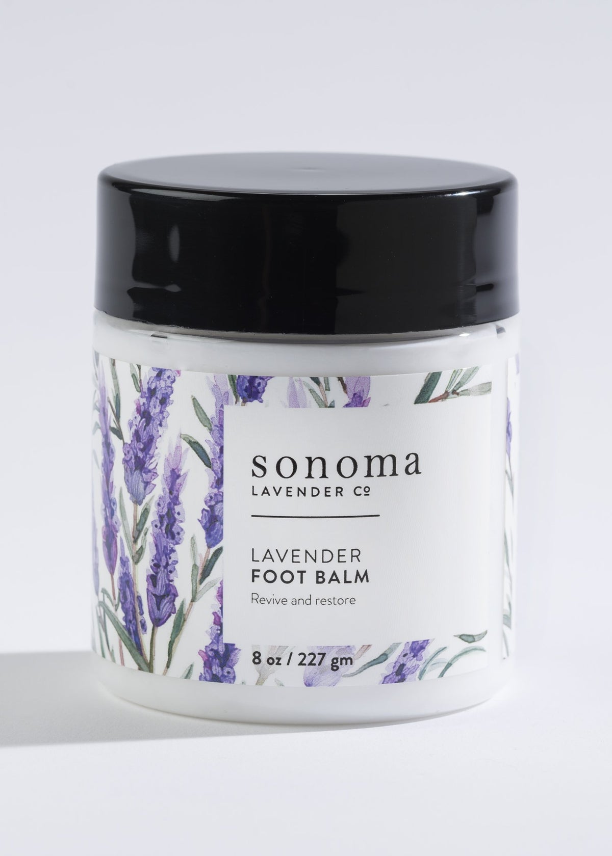 A jar of Sonoma Lavender Foot Balm labeled "lavender foot balm, revive and restore," featuring lavender illustrations and enriched with Lavender Essential Oil, displayed against a white background.