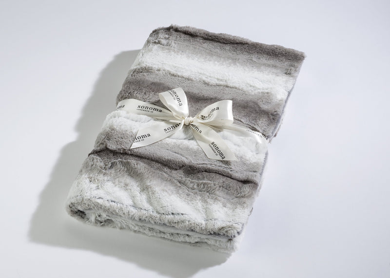A plush gray Sonoma Lavender Angora Platinum SPA BLANKIE neatly folded and tied with a white ribbon labeled "sonoma" rests against a white background. The texture appears soft and fluffy, infused with aromatic heat for muscle-relaxation.