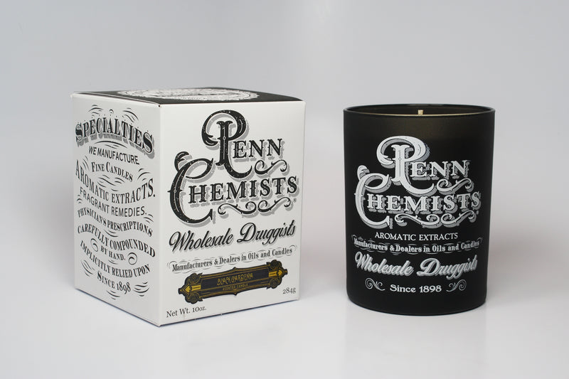 A Penn Chemists Classic Candle - Black Gardenia with "penn chemists" label next to its matching white box featuring decorative typography and vintage designs, set against a plain white background, exudes the enchanting aroma of Tahitian