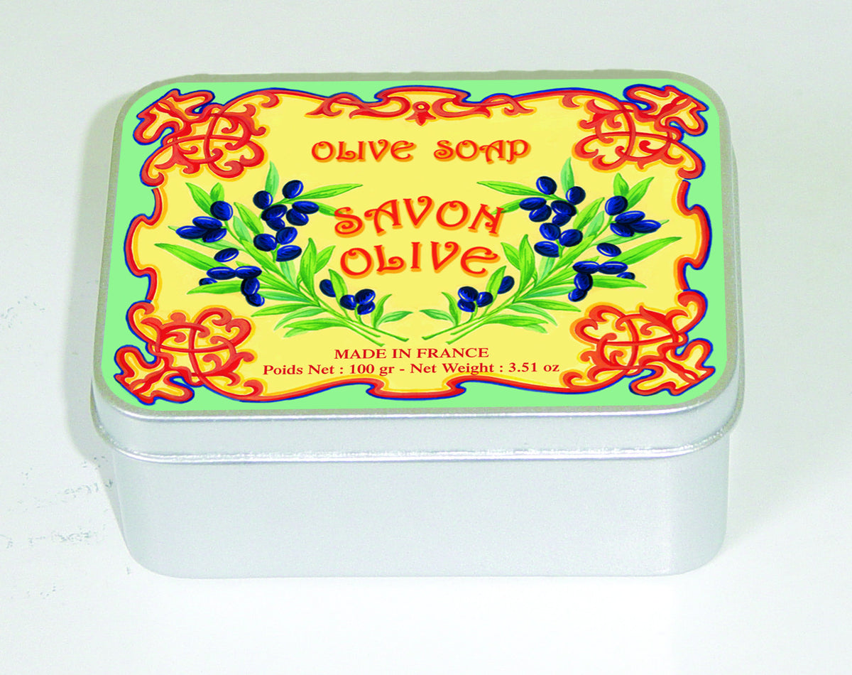A colorful tin of Le Blanc Olive 100gm Soap Tin labeled "savon d'olive," with ornate blue and yellow patterns and olive branch illustrations. Text indicates it is made in Provence, with a weight of