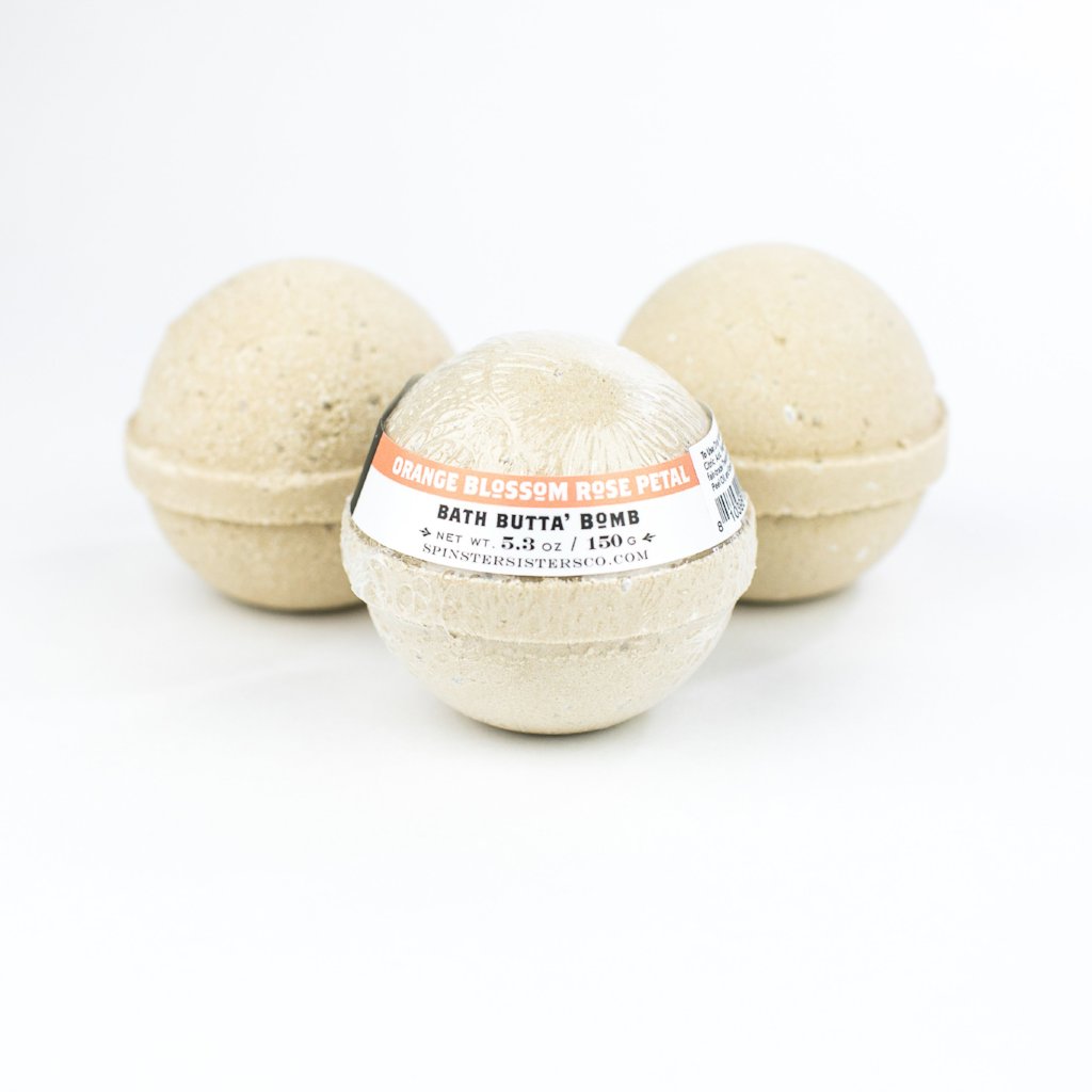 Three beige Spinster Sisters Bath Butta' Bombs enriched with shea butter, with a label on the front bath bomb reading "orange blossom rose petal bath butter bomb," displayed against a white background.