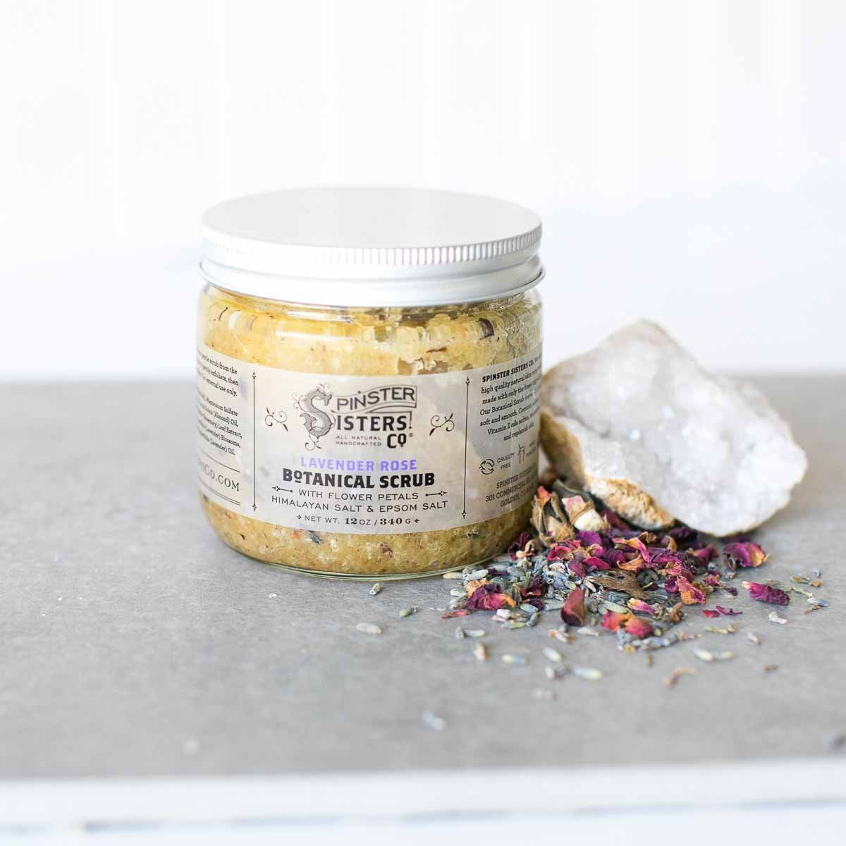 A jar of Spinster Sisters Lavender Rose Botanical Scrub on a light background, with scattered dried flowers and crystal beside it. The label lists natural, moisturizing ingredients.