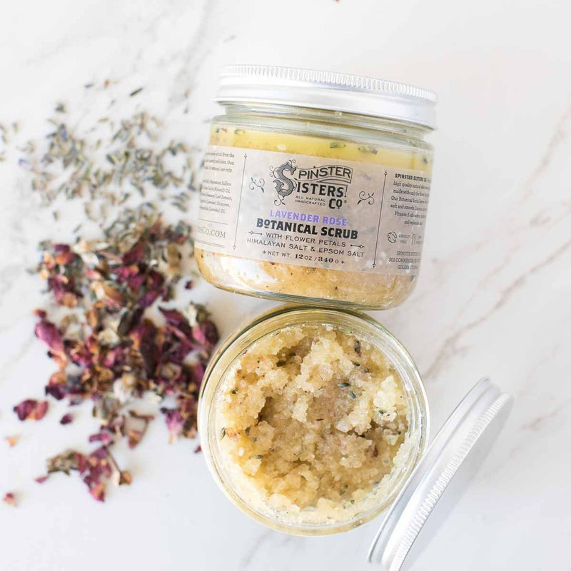 Two jars of Spinster Sisters, Co. Lavender Rose Botanical Scrub on a marble surface, one open with product visible, surrounded by scattered dried flowers.