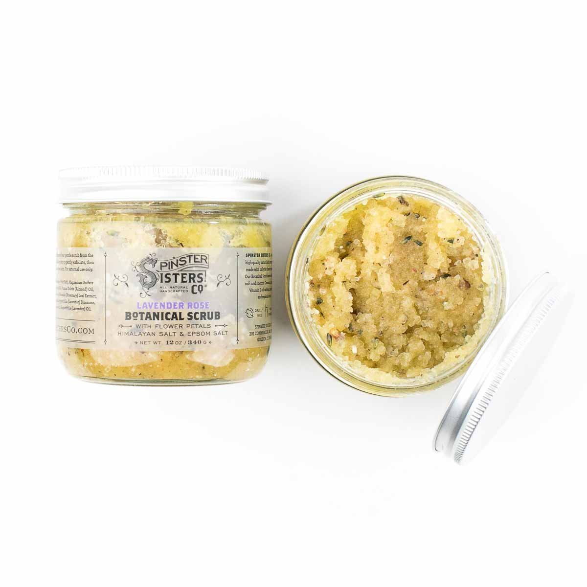 A jar of Spinster Sisters Lavender Rose Botanical Scrub alongside an open container of the moisturizing scrub with a small scoop, displayed on a white background.