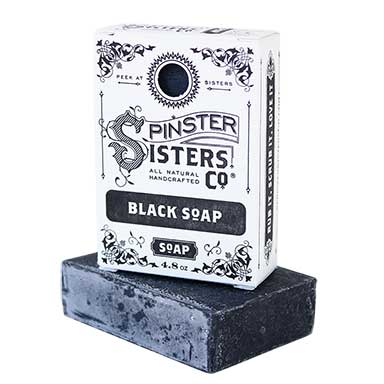 A black bar of Spinster Sisters Black Soap w/ Activated Charcoal (Coriander Lemon) is displayed in front of its biodegradable packaging. The box, labelled "Spinster Sisters, Co. All Natural Handcrafted Co. Black Soap," features ornate black and white design.