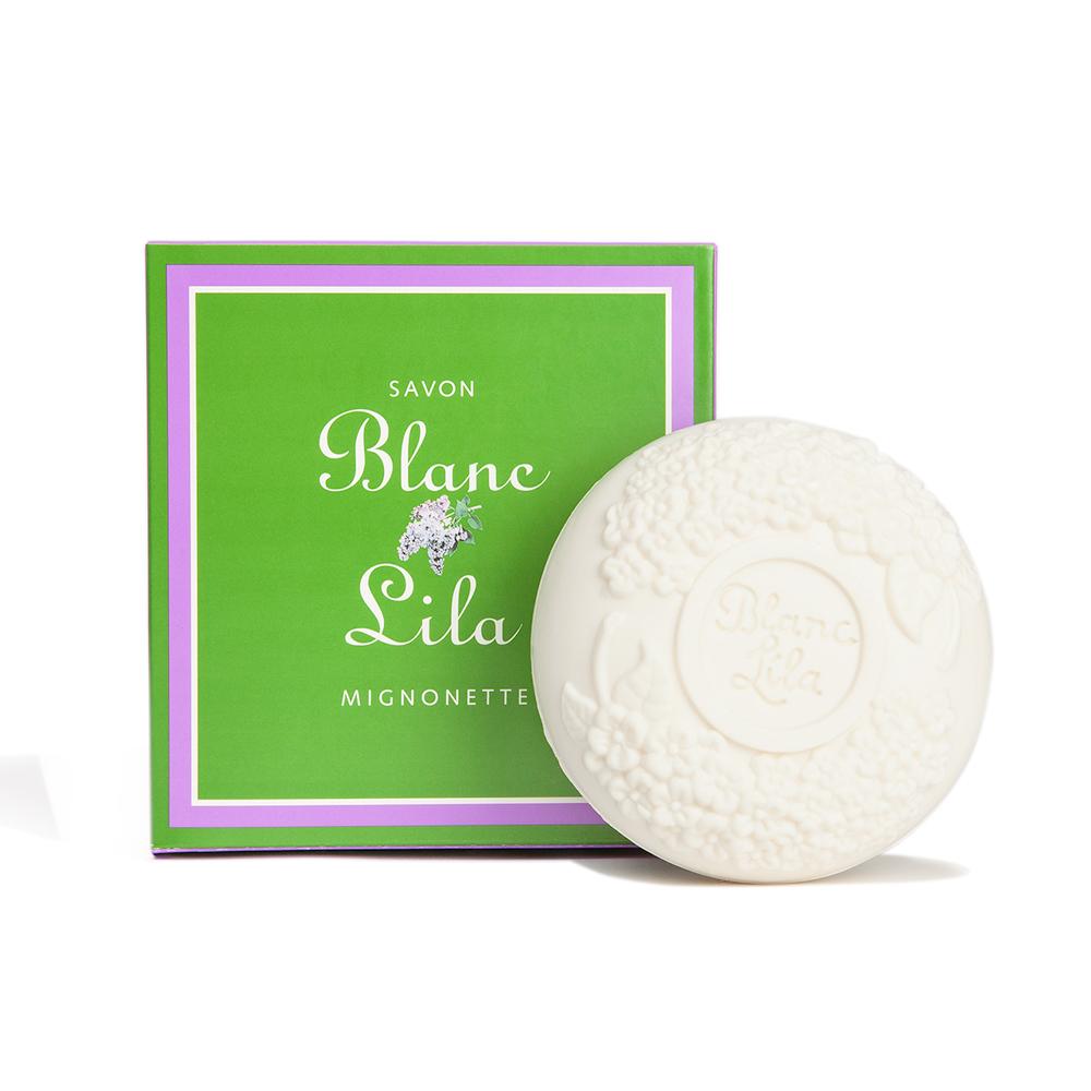 A bath bar of Blanc Lila (White Lilac) Bathing Bar soap from Soaps & Soaks From Around The World with embossed floral designs next to its green and purple packaging labeled "savon blanc lila mignonette," imported from Belgium.