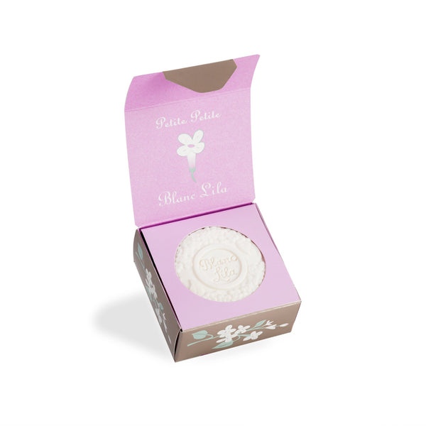 A decorative open box with a brown exterior featuring floral designs and a light pink interior holds a lilac bar embossed with "Blanc Lila." Inside, the white round soap radiates elegance, perfectly complementing the charm of Kala's Blanc Lila Petite Petite treasure.