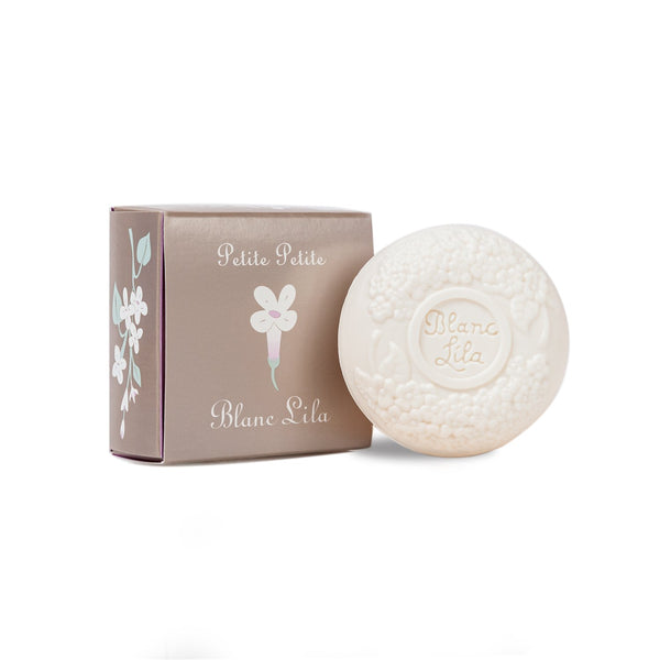 A round, white soap adorned with embossed floral designs rests beside a gray box. The packaging features an elegant illustration of white and pink flowers, along with the text "Blanc Lila Petite Petite" by Kala, capturing the essence of this lilac bar beautifully.