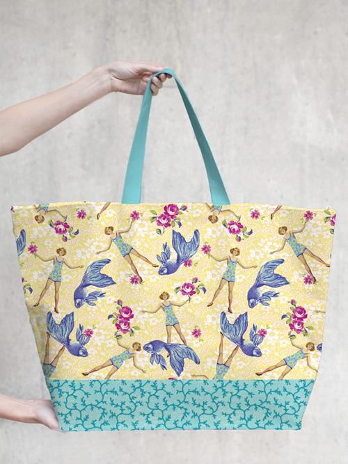 A person holding a large Margot Elena tote bag with a colorful TokyoMilk Blue Goldfish design featuring stylized birds and floral patterns on a grey background.