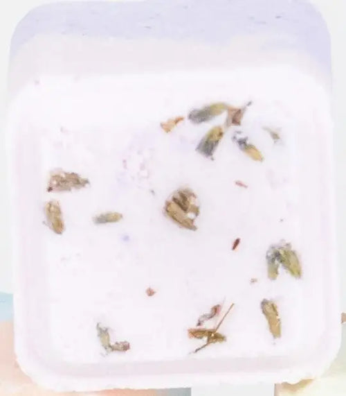 A Lizush Lavender Shower Steamer, filled with a creamy substance, sprinkled with small pieces of nuts and herbs composed of natural ingredients, set against a light, blurry background.