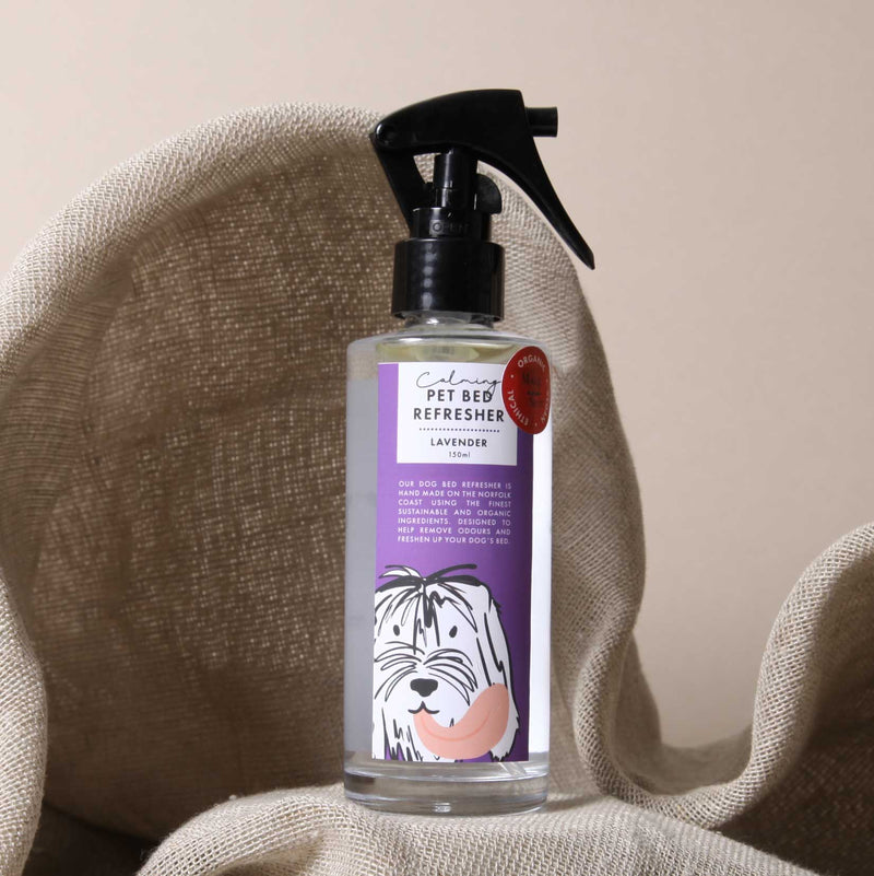 A clear spray bottle labeled "Made & Sent Calming Dog Bed Refresher lavender" with an illustration of a cute, shaggy dog on the label, set against a beige cloth backdrop, contains essential oils.