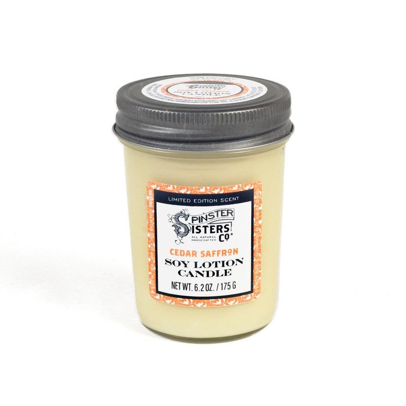 A Spinster Sisters Soy Candle - Cedar Saffron in a clear glass jar, labeled "cedar saffron" from "Spinster Sisters," with an orange and white label. This aromatherapy massage candle is a creamy