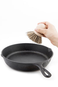 A person's hand cleans a cast iron skillet with an Andrée Jardin Tradition Saucepan Brush against a white background.