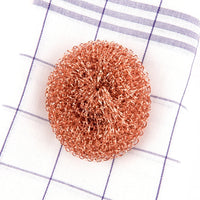 A Andrée Jardin Tradition Copper Scrubber lying on a blue and white checkered cloth. The pad is circular and made of intertwined metallic fibers, giving a textured appearance.