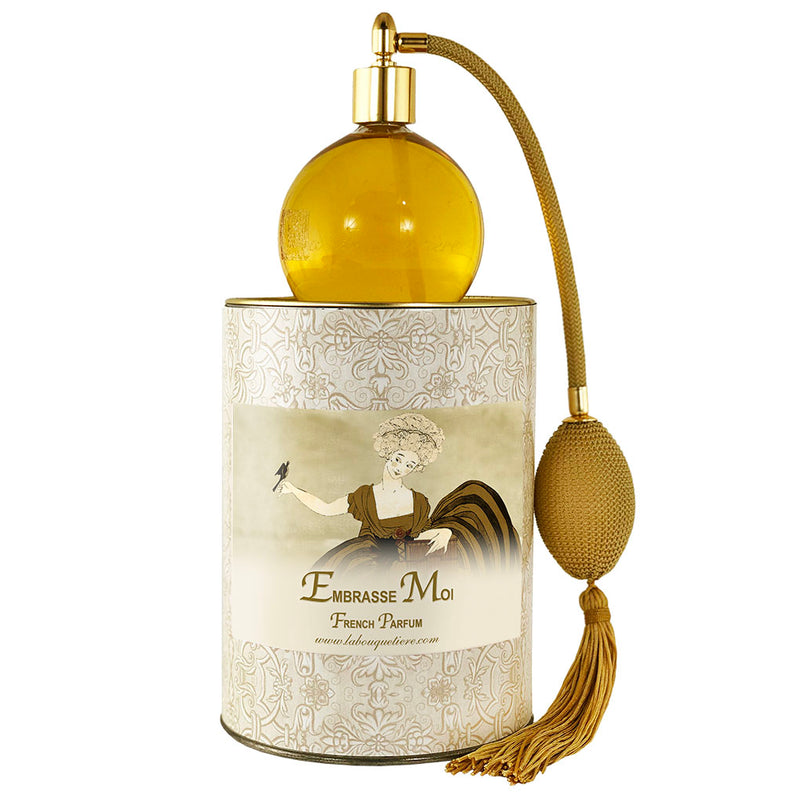 A vintage La Bouquetiere Embrasse Moi French Perfume bottle with a spherical golden top and tassel, housed in an ornate cylindrical container featuring a classic illustration and text 'embrasse moi french parfum'.