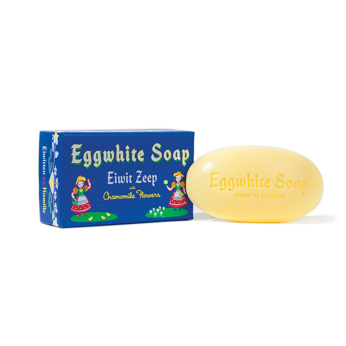 A bar of Eggwhite Facial Single Soap - Belgium is displayed in front of its packaging. The soap, a light yellow color with "Eggwhite Soap - Made in Belgium" inscribed, boasts soothing chamomile. The blue box features the text "Eggwhite Facial Single Soap - Belgium - Eiwit Zeep - Chamomile Flowers" and colorful illustrations, epitomizing Belgian beauty bars from Soaps, Soaks & Pampering Products From Around the World.