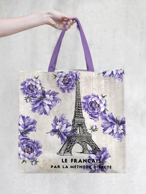 A person's hand holding a TokyoMilk Tote Bag - French Kiss Market Tote featuring a design with the Eiffel Tower, purple flowers, and French text on a light background by Margot Elena.