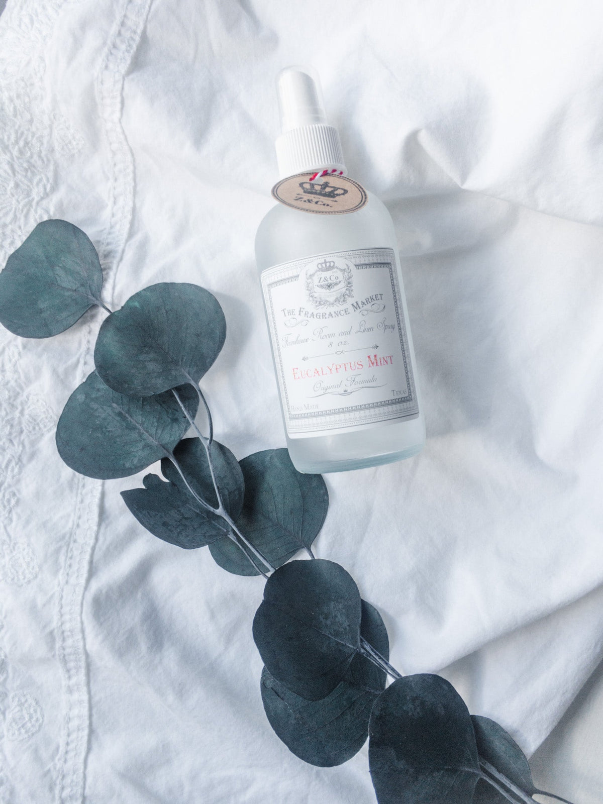 A bottle of Z&Co. Eucalyptus Mint Farmhouse Room & Linen Spray sits on a white fabric background, accompanied by a sprig of eucalyptus leaves. The setting is soft.